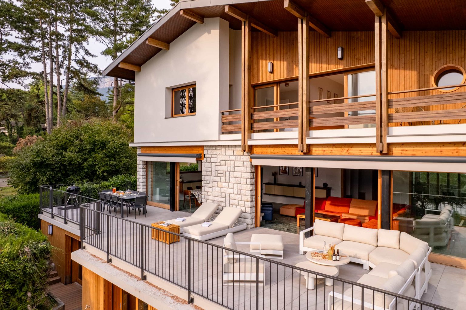 Façade en bois et pierre de la Villa Sarmaz avec sa grande terrasse meublée, au milieu des arbres